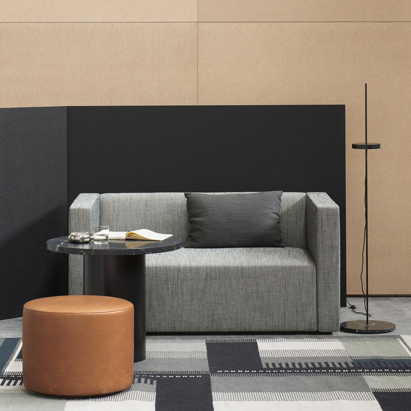 A modern living room includes a gray sofa, a black side table with a book and glasses, and a round brown ottoman. A sleek floor lamp stands nearby. The geometric-patterned carpet complements the black and tan walls. E15s CT09 ENOKI tables enhance the space with their rich marble tops and solid wood bases.