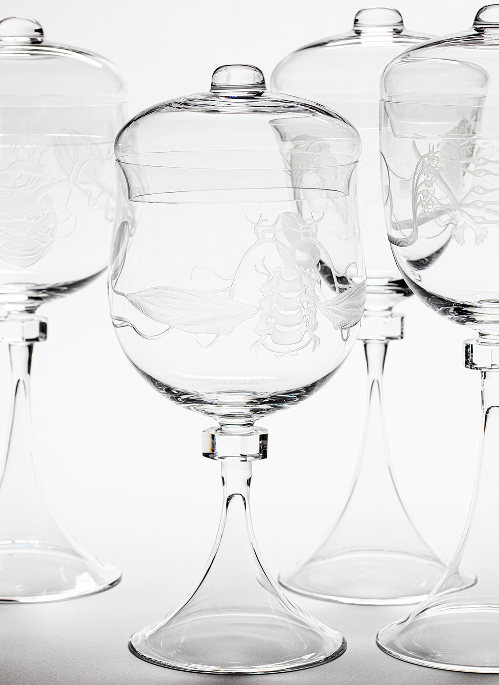 A close-up of Lobmeyrs Goblets of Humanity showcases tall, clear glass vessels with lids, copper wheel engravings of lobsters and fish, lead-free crystal construction, elegant elongated stems, and a tight arrangement against a plain background.