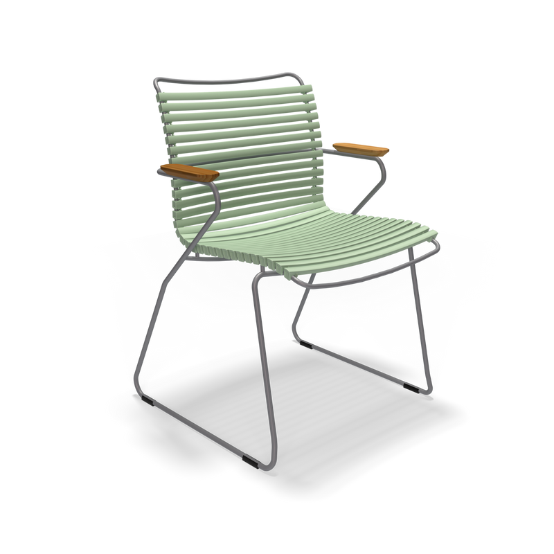 The Click Dining Chair with Armrest by Houe features modern green slatted plastic lamellas, a thin metal frame, curved wooden armrests, and sled-style legs. Its weather-resistant design is ideal for outdoor use and offers a minimalist look against a plain black background.