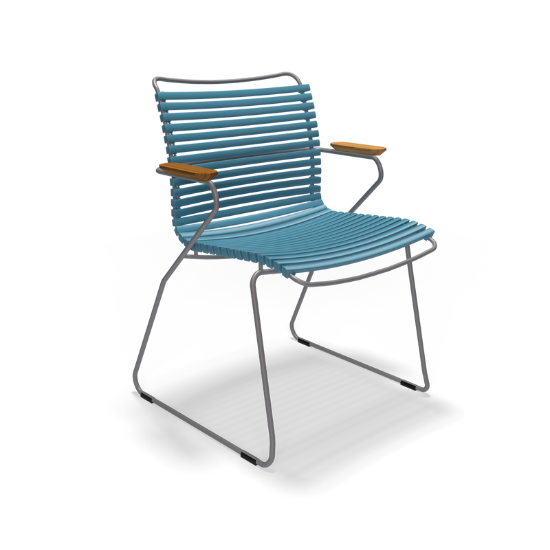 The Click Dining Chair with Armrest by Houe is a stylish, modern outdoor chair featuring weather-resistant blue plastic lamellas for the slatted seat and backrest, complemented by thin metal legs and wooden armrests.