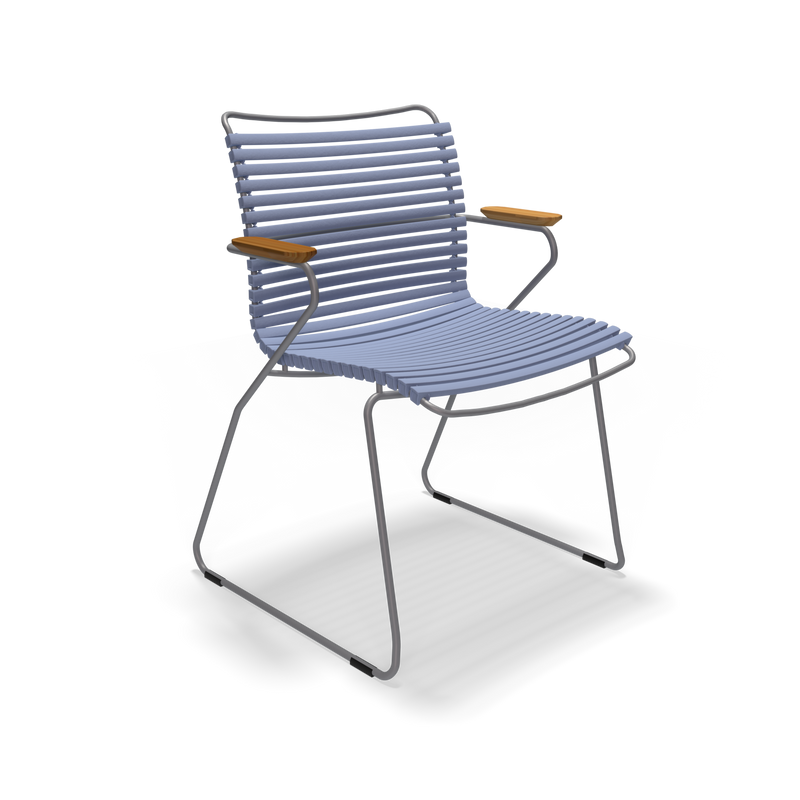 The Click Dining Chair with Armrest by Houe is a modern, minimalist chair with a ribbed blue seat and backrest, metal frame, and wooden armrests. Its weather-resistant design makes it ideal for outdoor use, featuring a sleek sled base against a black background.