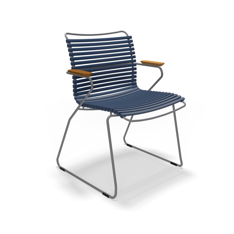 The Click Dining Chair with Armrest by Houe features a sleek design with weather-resistant, ribbed dark blue plastic lamellas for the seat and backrest, complemented by thin metal legs and wooden armrests. The minimalist and contemporary look is set against a plain black background.