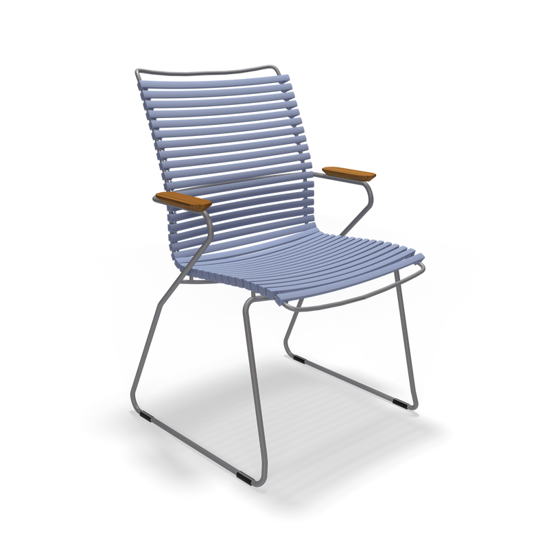 The Click Dining Chair with Armrest - Tall Back by Houe is a modern, minimalist outdoor chair featuring a curved, slatted blue seat and backrest made from UV-protected, weather-resistant material, slender metal legs, wooden armrests, and set against a black background.