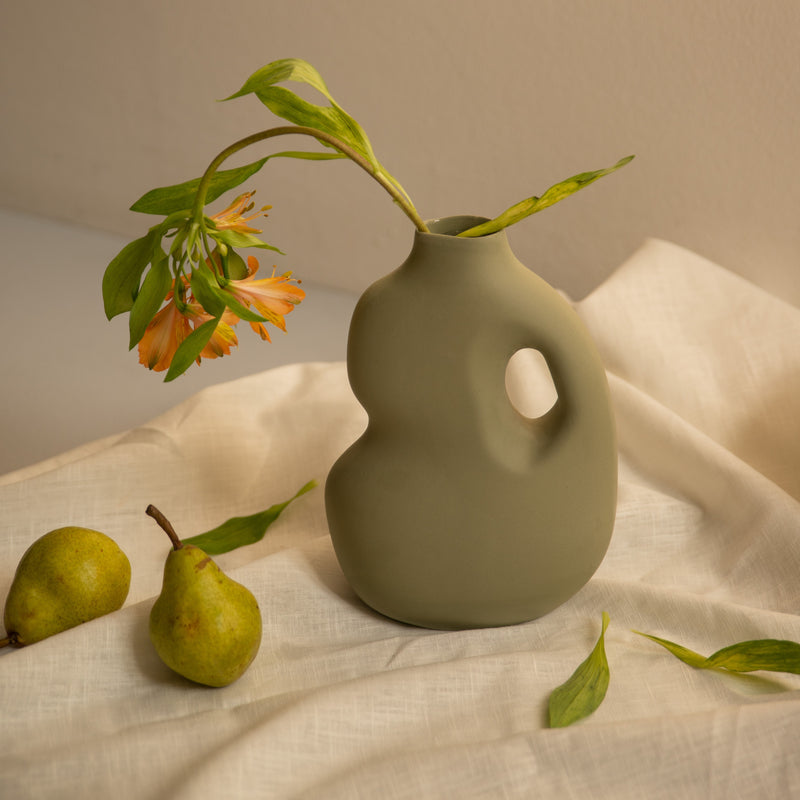 The Schneids Aura II, a matte green ceramic vase with a unique curvy design, holds an orange stem of flowers with long green leaves. Two pears rest nearby on light, textured fabric with scattered leaves, creating a minimalist and natural ambiance.