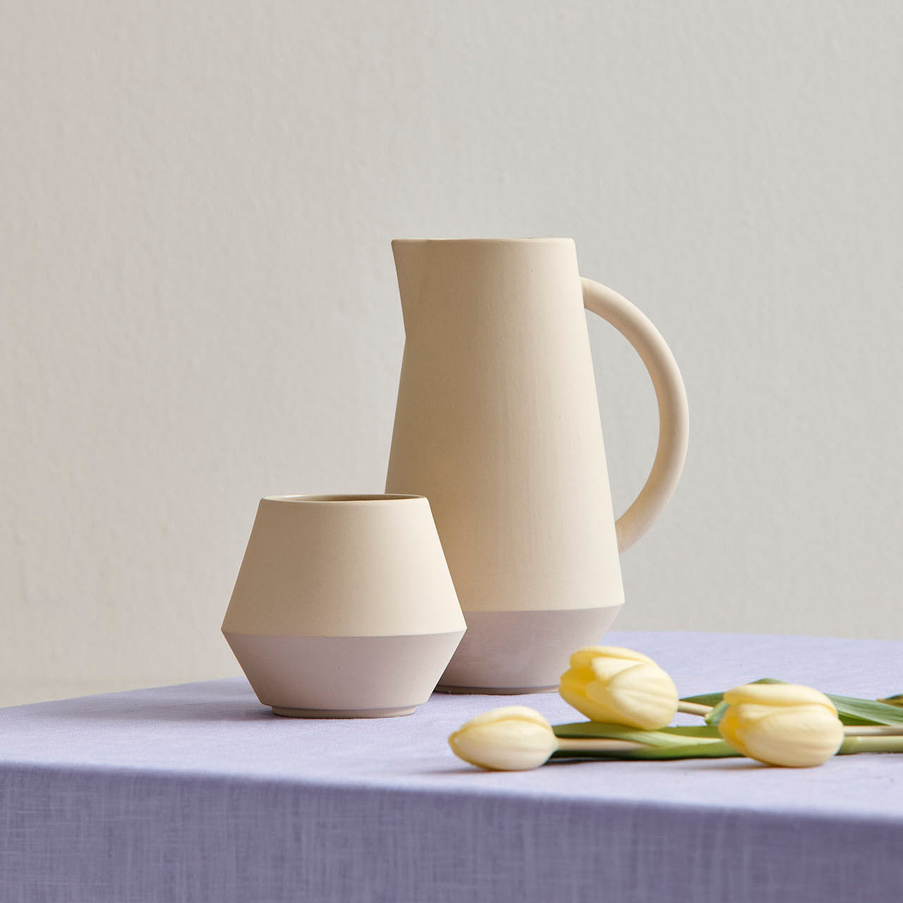 A cream Unison Sugar Bowl by Schneid, adorned with geometric designs, sits on a light purple tablecloth alongside a matching cup. Three yellow tulips lie beside this elegant set. A pale, neutral wall in the background accentuates the handcrafted colors of the ceramic tableware.