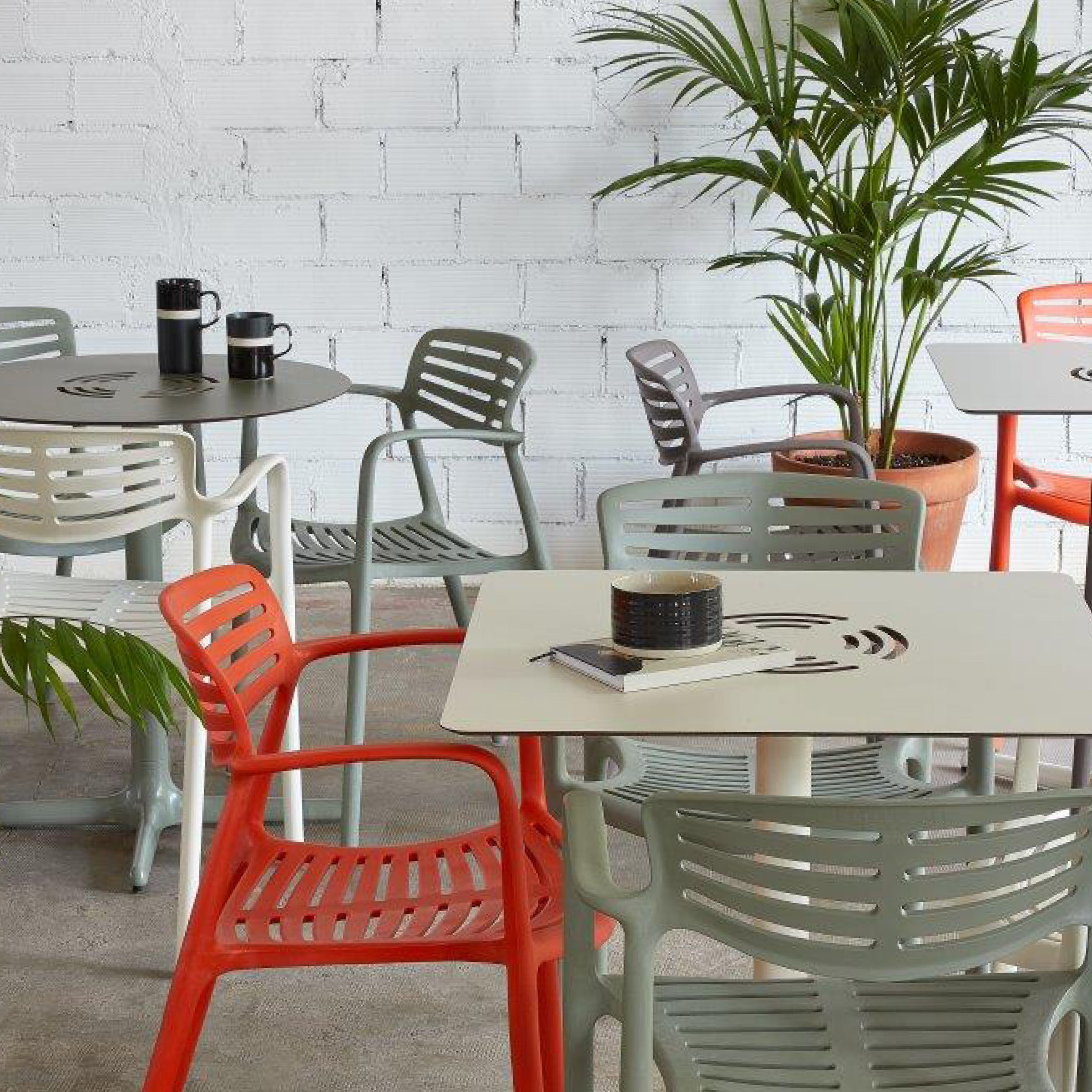 In a modern café, Resols Toledo Aire Armchairs add vibrant flair with red and gray seating around sleek metal tables. Potted plants grace the background, and coffee cups rest on the tables, enhancing the cozy ambiance.