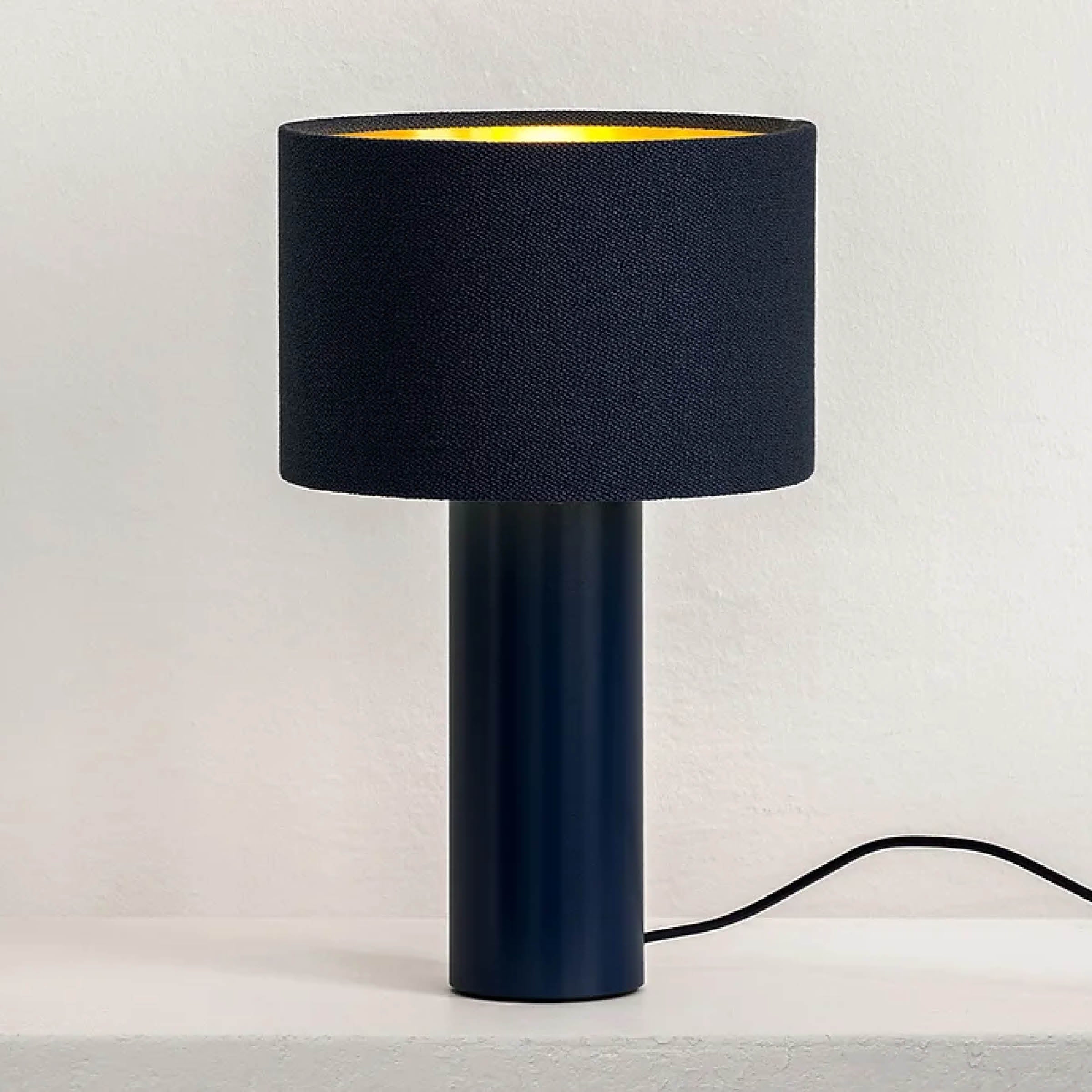 The ALL ROUND MINI Table Lamp by Victor Foxtrot features a black cylindrical base and a wool drum shade, creating a warm glow. Situated on a white surface against a light wall, its black power cord extends from the base.