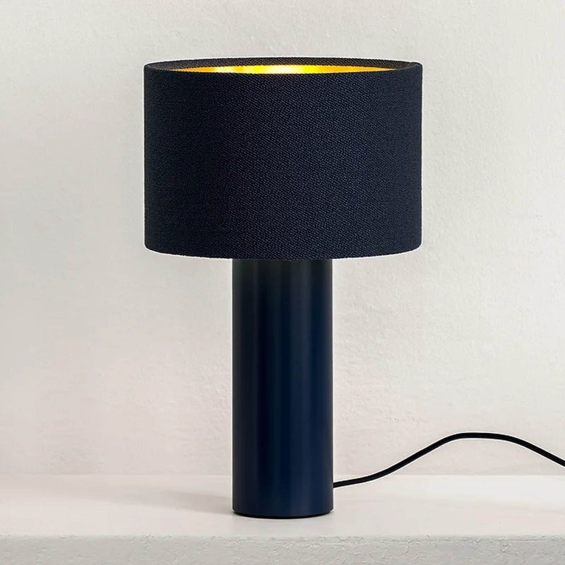 The ALL ROUND MINI Table Lamp by Victor Foxtrot features a black cylindrical base and a wool drum shade, creating a warm glow. Situated on a white surface against a light wall, its black power cord extends from the base.