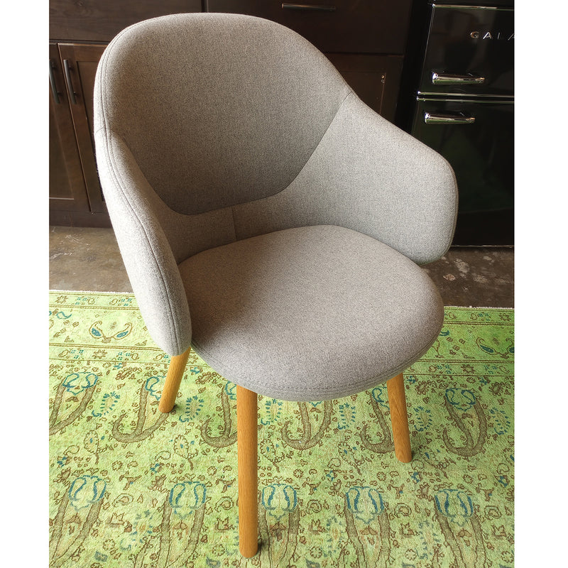 The Armchair Albu by TON, in modern gray upholstery with a curved backrest and wooden legs, sits on a green carpet with a paisley pattern. In the background are dark wooden cabinets and the base of a black appliance.