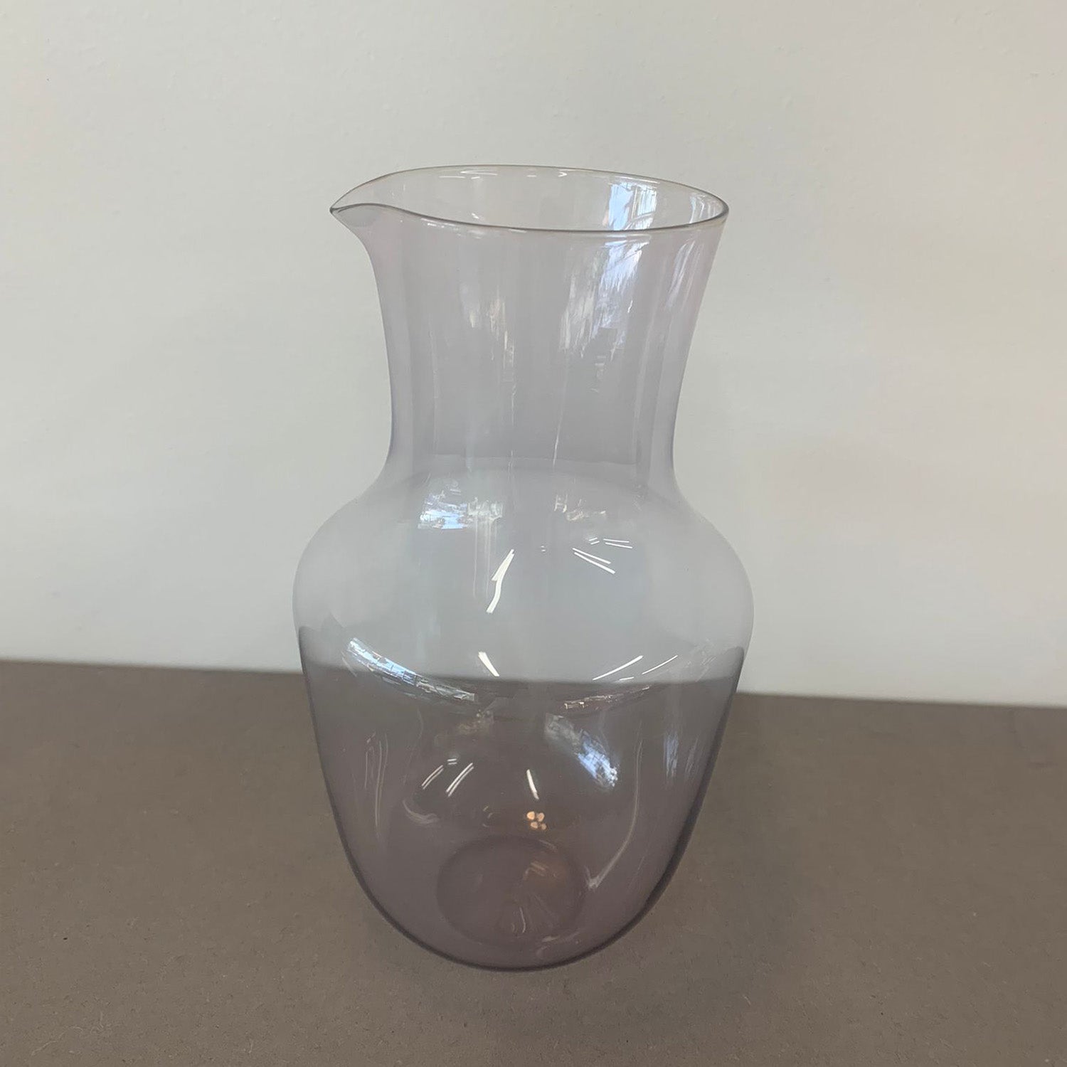 The Lobmeyr Alpha Water Pitcher - Sale is a translucent lavender glass container with a stackable crystal-like appearance, subtly fading from clear at the top to purple at the bottom, set on a gray surface against a light gray backdrop for an elegant touch.