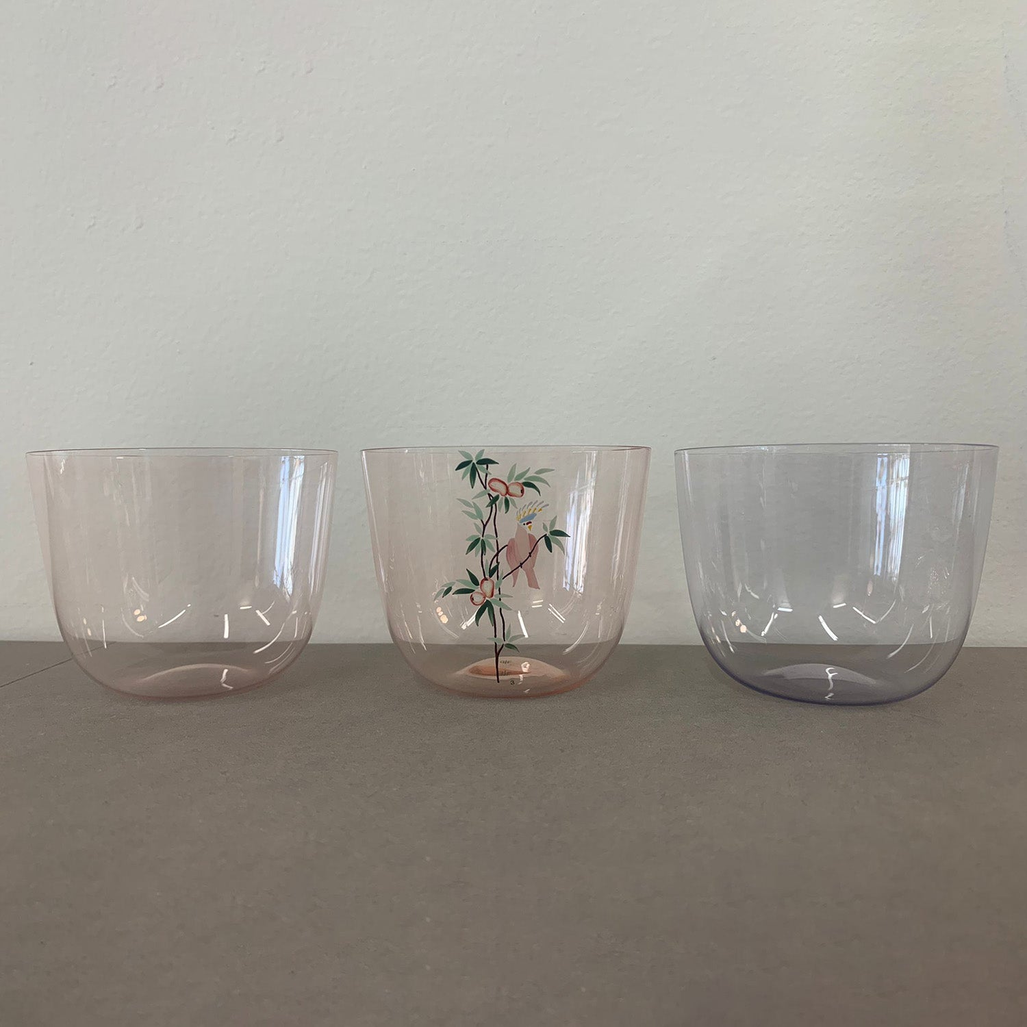 Three transparent glass cups sit on a flat surface against an off-white wall. The left one, the Alpha Water Tumbler by Lobmeyr, is slightly pink. The middle cup showcases a bamboo design, and the right is light gray. Each piece is expertly crafted from mouth-blown lead-free crystal.