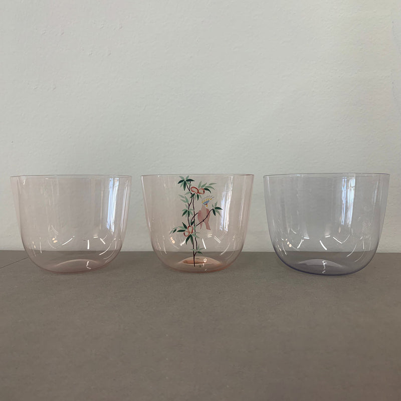 Three transparent glass cups sit on a flat surface against an off-white wall. The left one, the Alpha Water Tumbler by Lobmeyr, is slightly pink. The middle cup showcases a bamboo design, and the right is light gray. Each piece is expertly crafted from mouth-blown lead-free crystal.