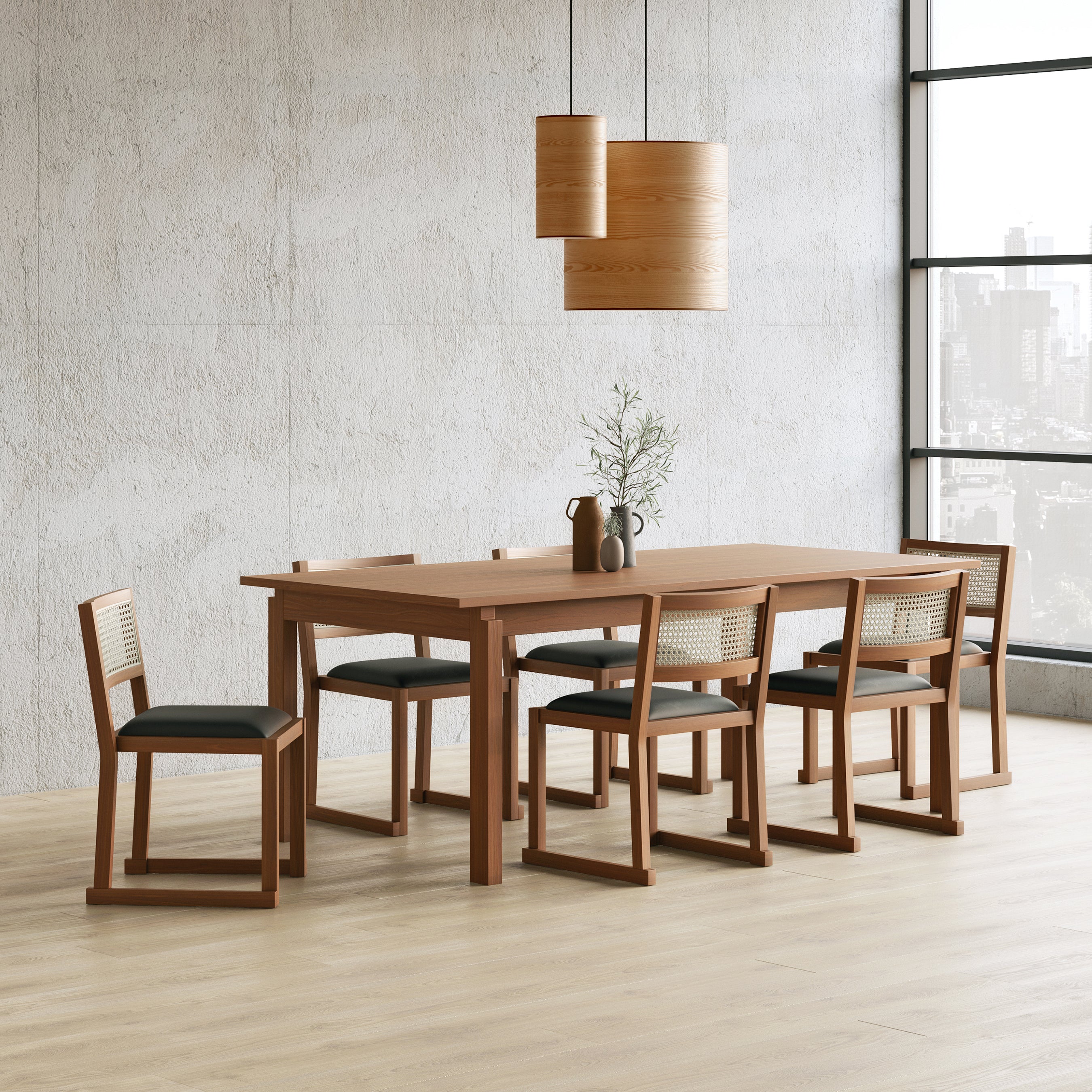 A modern dining room features a Gus Modern Annex Extendable Dining Table, six cane-back chairs with black cushions, a brown vase with greenery, two wooden pendant lights, and large windows overlooking the city.