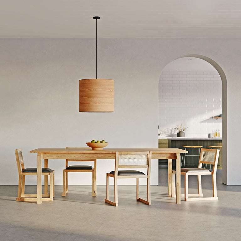 A modern dining room features a Gus Modern Annex Extendable Dining Table, four matching chairs, a fruit bowl, and a wooden pendant light. An arched doorway opens to a kitchen with white tile walls and dark cabinets.