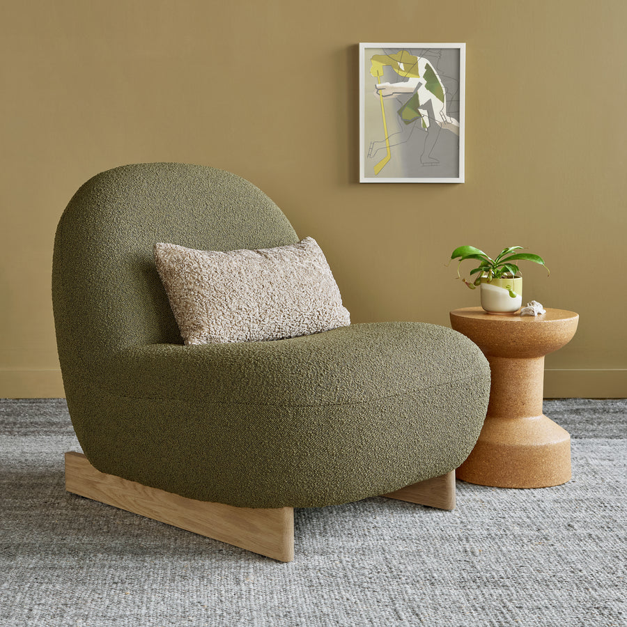 The Gus Modern Arlo Chair in olive green, paired with a beige textured pillow, sits by a small round table with a potted plant. Above, abstract art hangs on a tan wall over a gray rug for subtle Scandinavian flair.