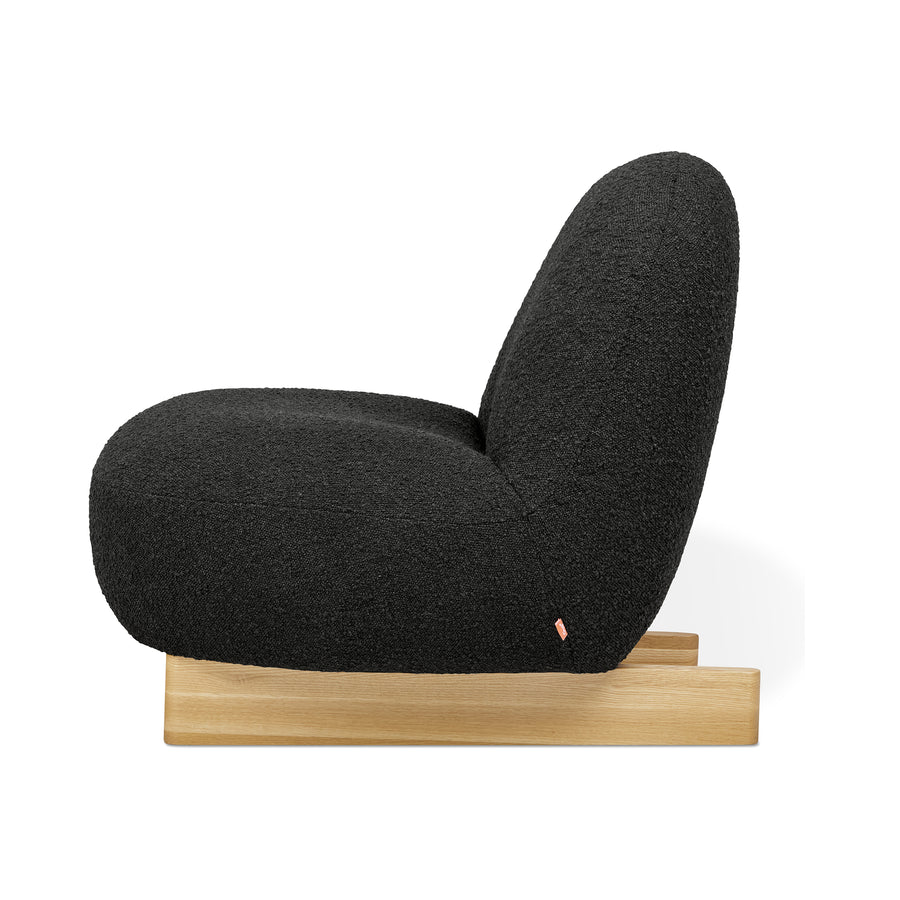 The Gus Modern Arlo Chair features a modern Scandinavian silhouette with thick, rounded lines, upholstered in rich black bouclé fabric on a low, light wood base and displayed from the side against a white background.