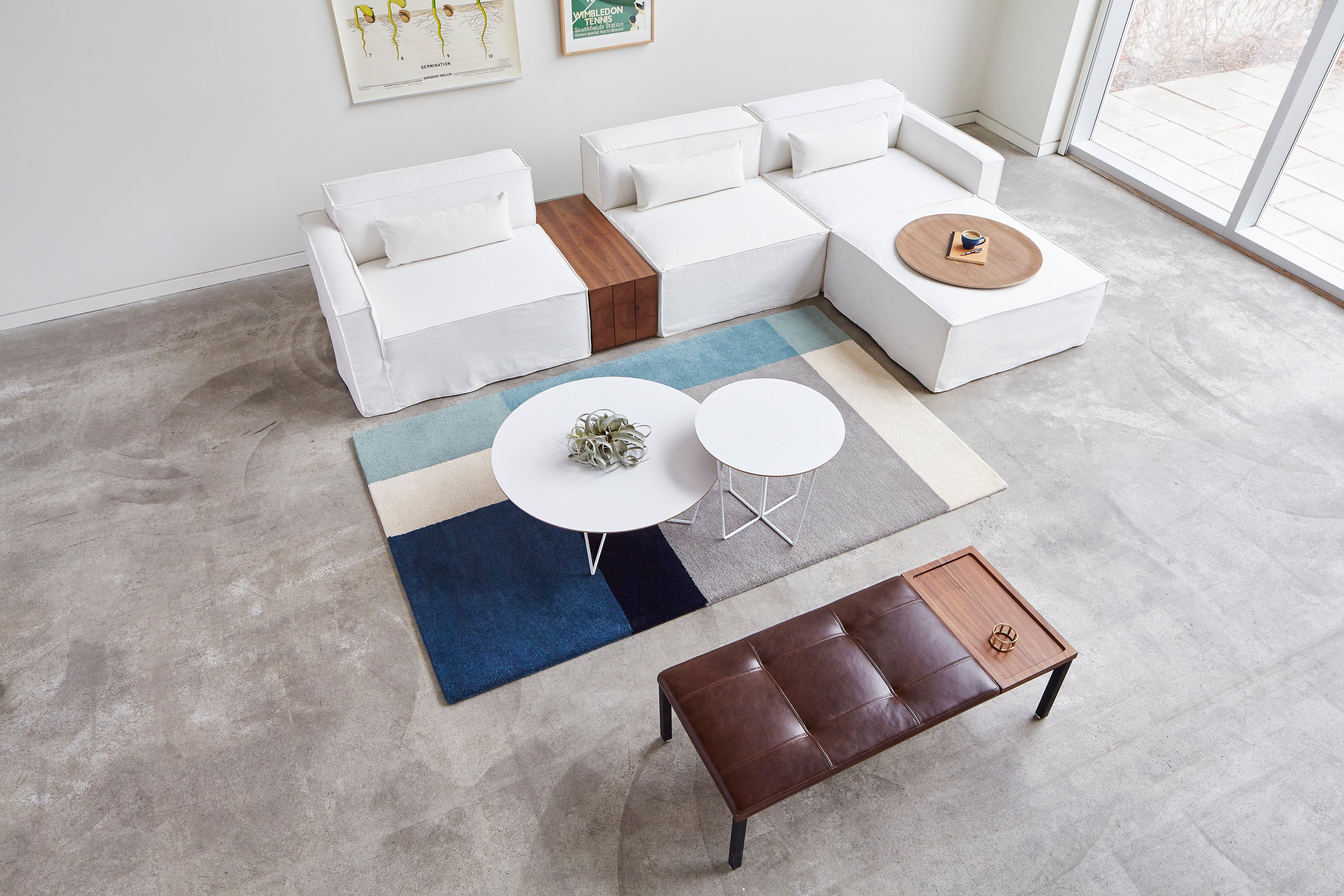 A modern living room with white sectional sofas, a brown leather bench, two Gus Modern Array End Tables with geometric bases, a blue and beige geometric rug, and large windows letting in natural light creates a contemporary accent.