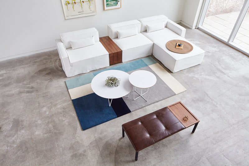 A modern living room with white sectional sofas, a brown leather bench, two Gus Modern Array End Tables with geometric bases, a blue and beige geometric rug, and large windows letting in natural light creates a contemporary accent.