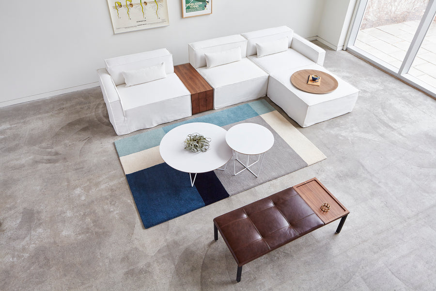 A modern living room with white sectional sofas, a brown leather bench, two Gus Modern Array End Tables with geometric bases, a blue and beige geometric rug, and large windows letting in natural light creates a contemporary accent.