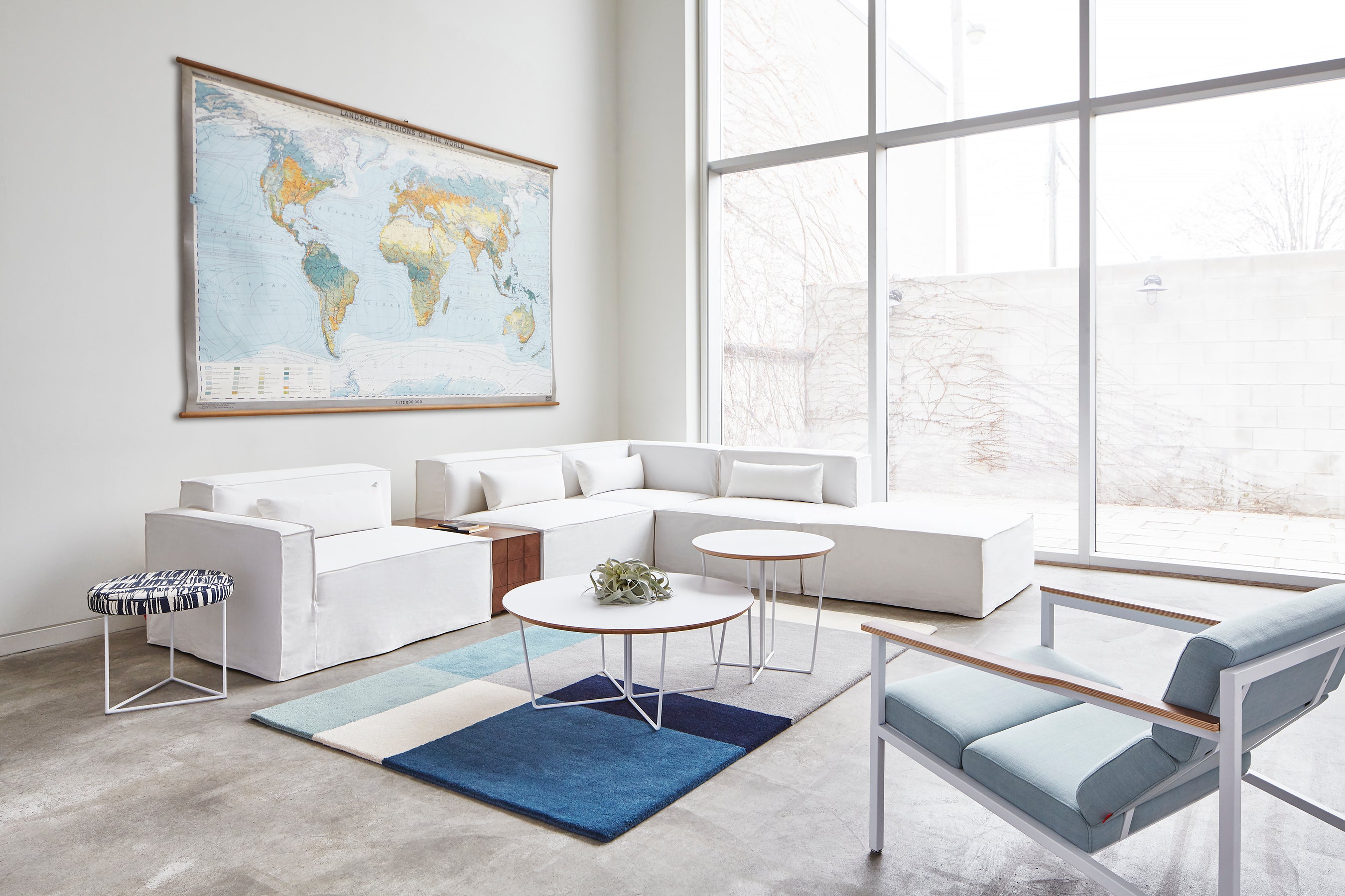 A modern, bright living room features white sectional sofas, a Gus Modern Array End Table, round geometric-base coffee tables, a blue geometric rug, a light armchair, and a world map. Floor-to-ceiling windows fill the space with natural light.