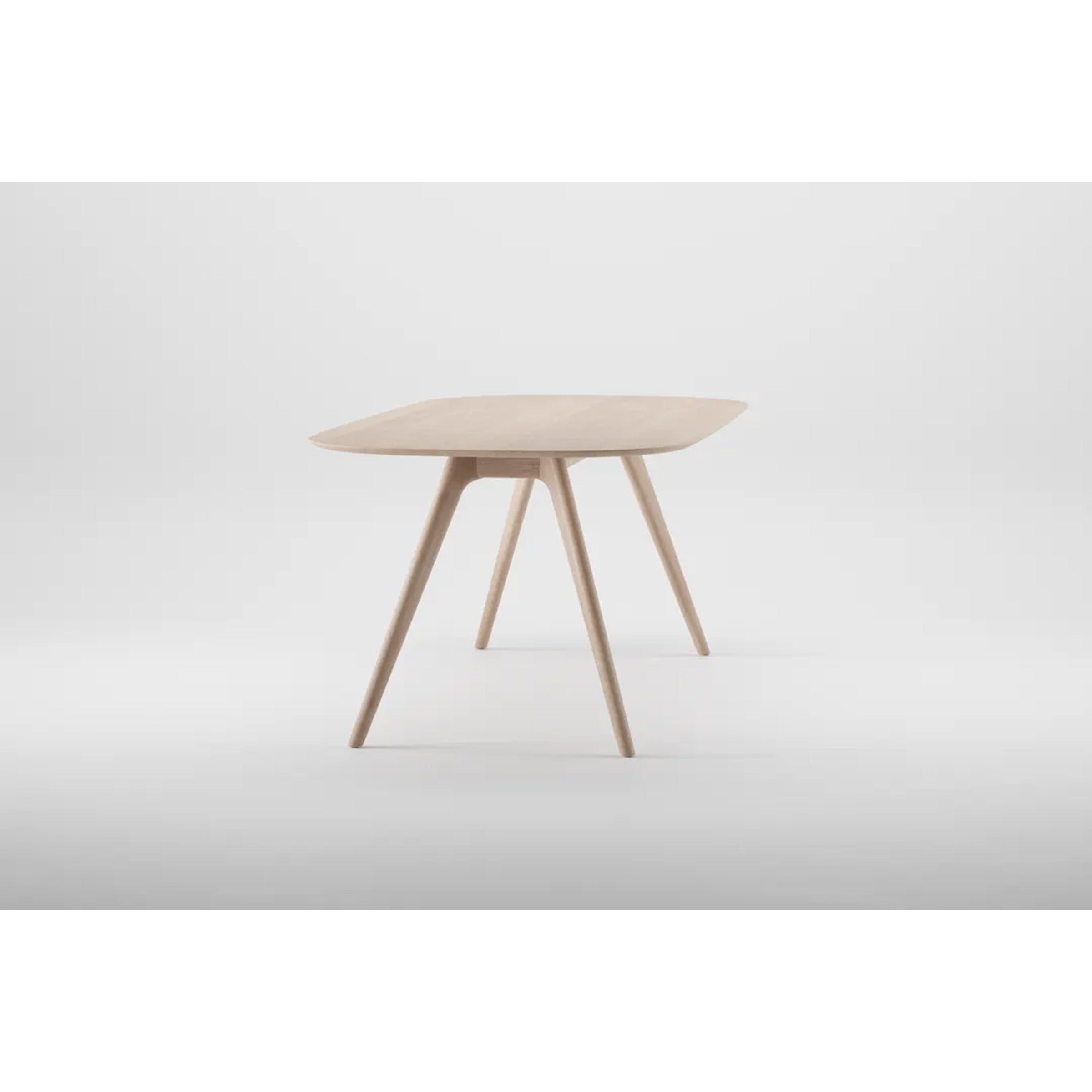 The COCO Table by Artisan is a sleek, contemporary wooden table featuring smooth curves and four tapered legs against a light gray background. Crafted from solid wood and finished with natural oils, it highlights environmentally-friendly materials.
