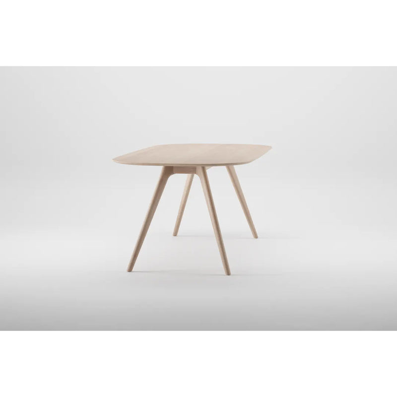 The COCO Table by Artisan is a sleek, contemporary wooden table featuring smooth curves and four tapered legs against a light gray background. Crafted from solid wood and finished with natural oils, it highlights environmentally-friendly materials.