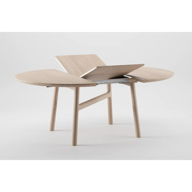 The DASH Extendable Table by Artisan is a solid wood round dining table with a minimalist design. Made from environmentally-friendly materials, it features an extendable leaf and four legs, set against a plain gray background, radiating elegance and sustainability.