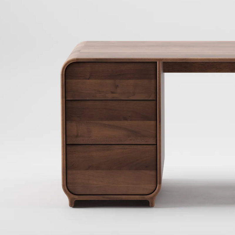 The ENY Working Desk by Artisan is a modern wooden desk with smooth, curved edges crafted from solid wood. It features three handle-less drawers on the left side, and the woods rich, natural finish is enhanced by natural oils against a plain white background.