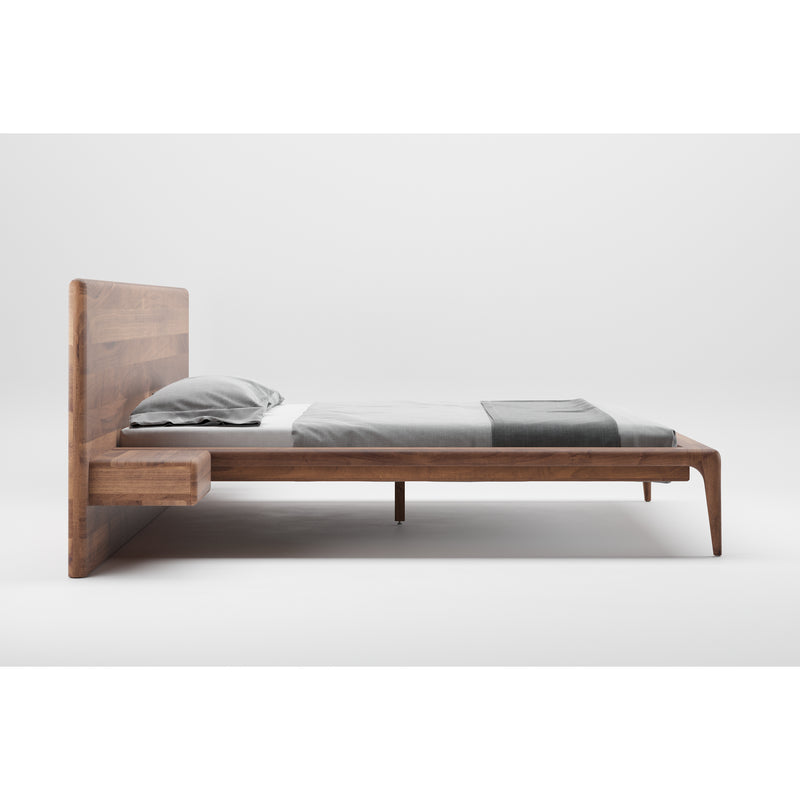 The LATUS Bed by Artisan features a minimalist design made from solid light brown wood with natural oil finish. It includes an attached side table and is shown with a gray duvet and pillows, set against a plain white background.