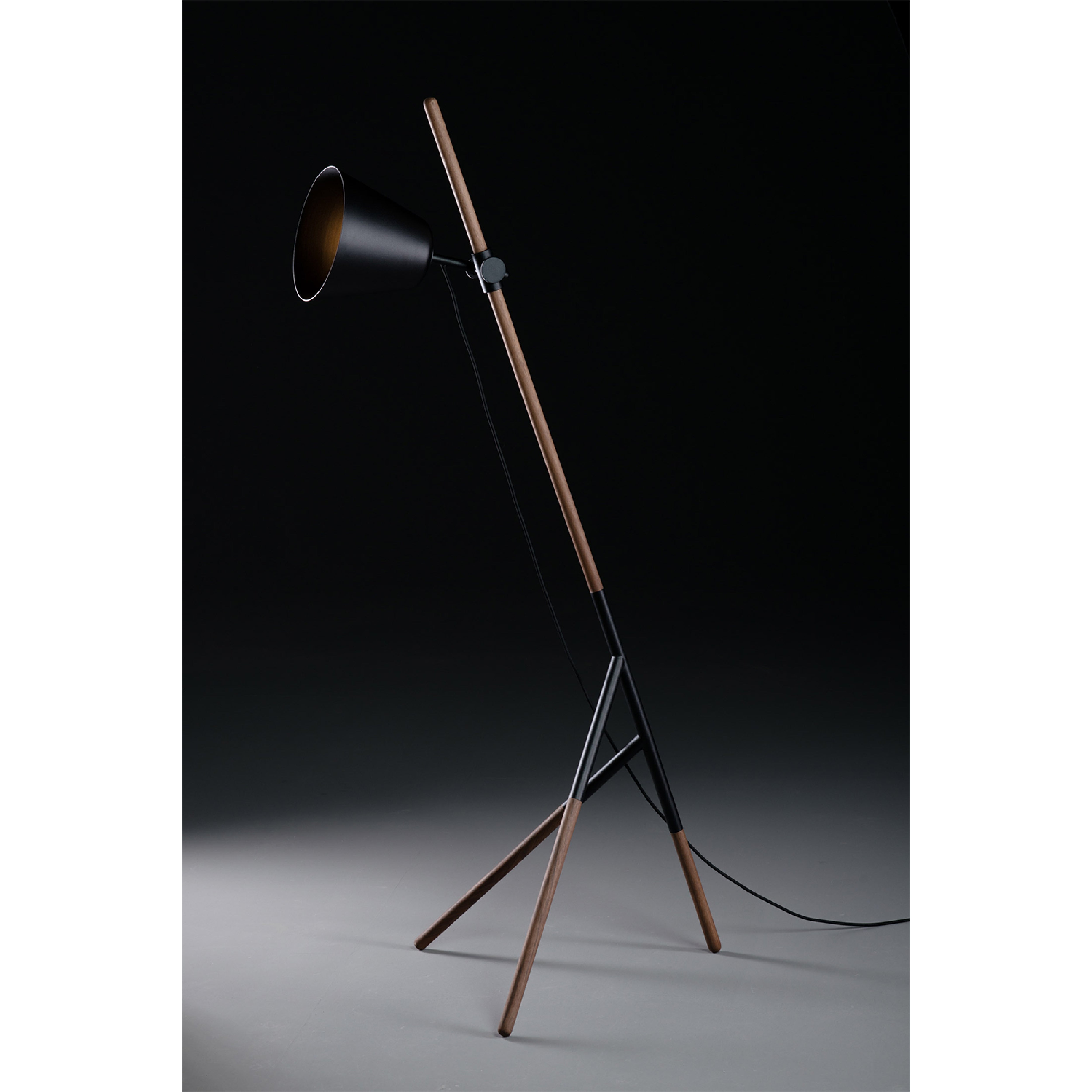 The Artisan INSERT Floor Lamp features a modern, minimalist design with a black conical shade on a solid wood tripod stand. This eco-friendly lamp leans at an angle, set against a dark backdrop to emphasize its sleek lines and contemporary aesthetic.