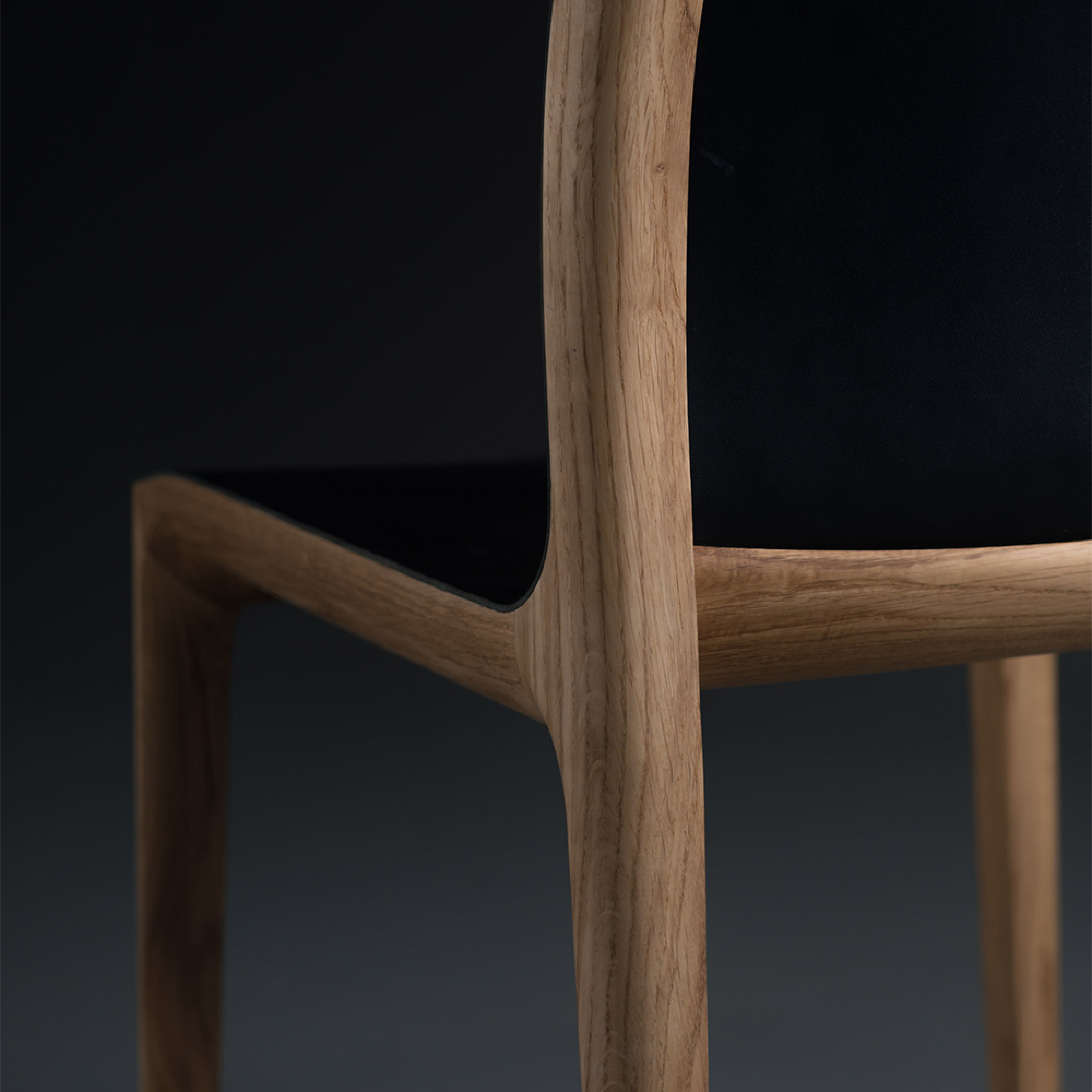 Close-up of the INVITO Chair by Artisan, featuring curved solid wood legs treated with natural oils and a smooth black seat. The dark background enhances its sleek, minimalist design crafted from eco-friendly materials.