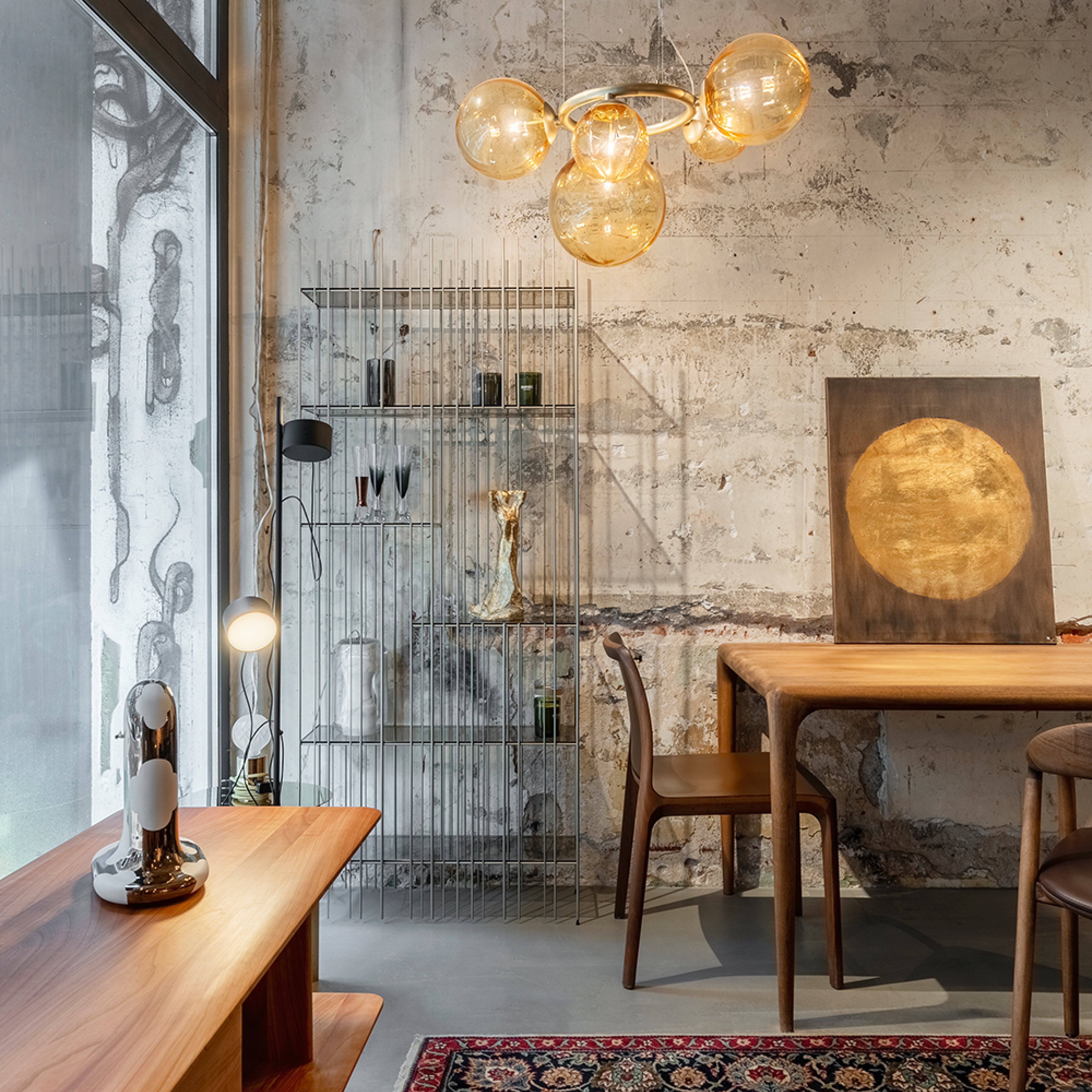 A modern room with rustic concrete walls includes an Artisan INVITO Chair alongside a solid wood table and bench. Decor items sit on a shelving unit, spherical lights from a chandelier hang above, natural light fills through a large window, and a painting with a gold circle leans against the wall.