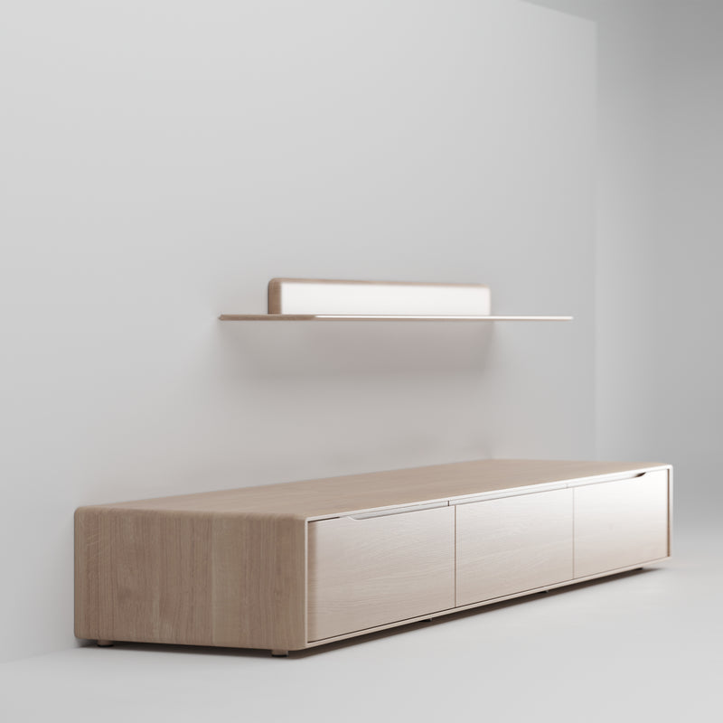 The Artisan INVITO MS Lowboard, a minimalist solid wood piece against a white wall, features three cabinet doors and a simple floating shelf above. Its sleek design and light wood tones with clean lines create a serene, contemporary look.