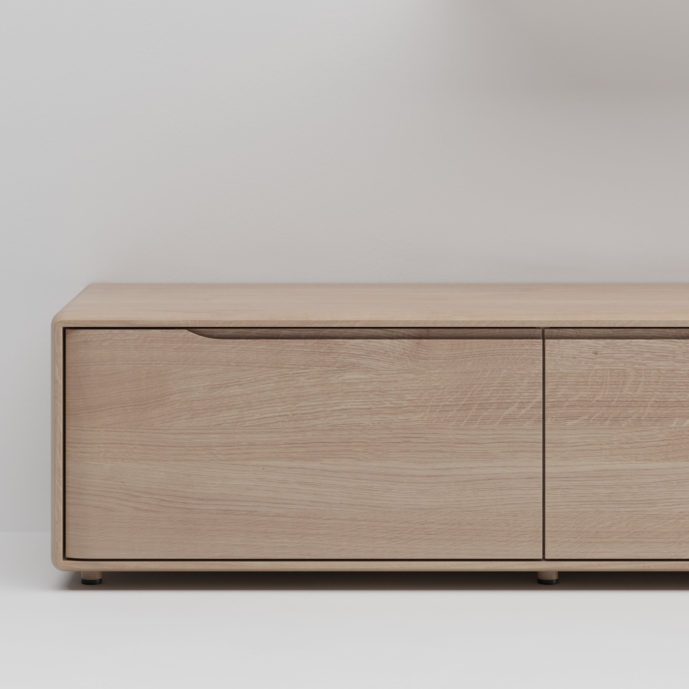 The Artisan INVITO MS Lowboard shines with its minimalist wooden design and light oak finish. Featuring two closed doors with subtle handles, it stands elegantly against a plain white wall and light floor, highlighting its craftsmanships simplicity.