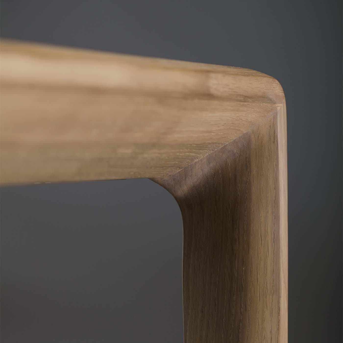 A close-up of the smooth, carved corner of the Artisans INVITO Table highlights its natural wood grain and elegant craftsmanship against a dark background.