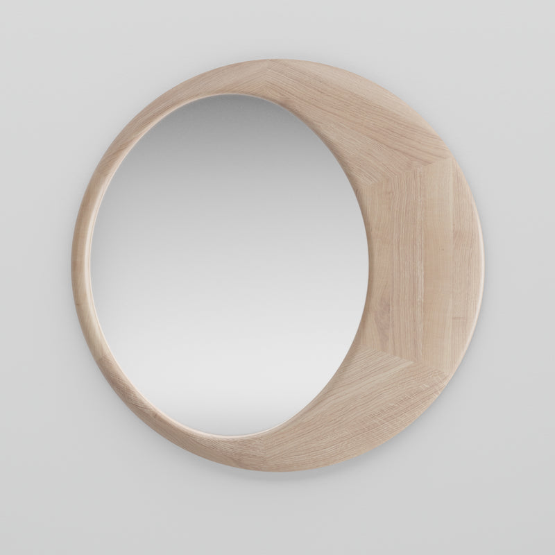 The Artisan Lana Mirror is circular, with a solid wood frame and unique design where the wood slightly overlaps the mirrors edge. Treated with natural oils, it offers a clear reflection against a white wall, made using eco-conscious materials.