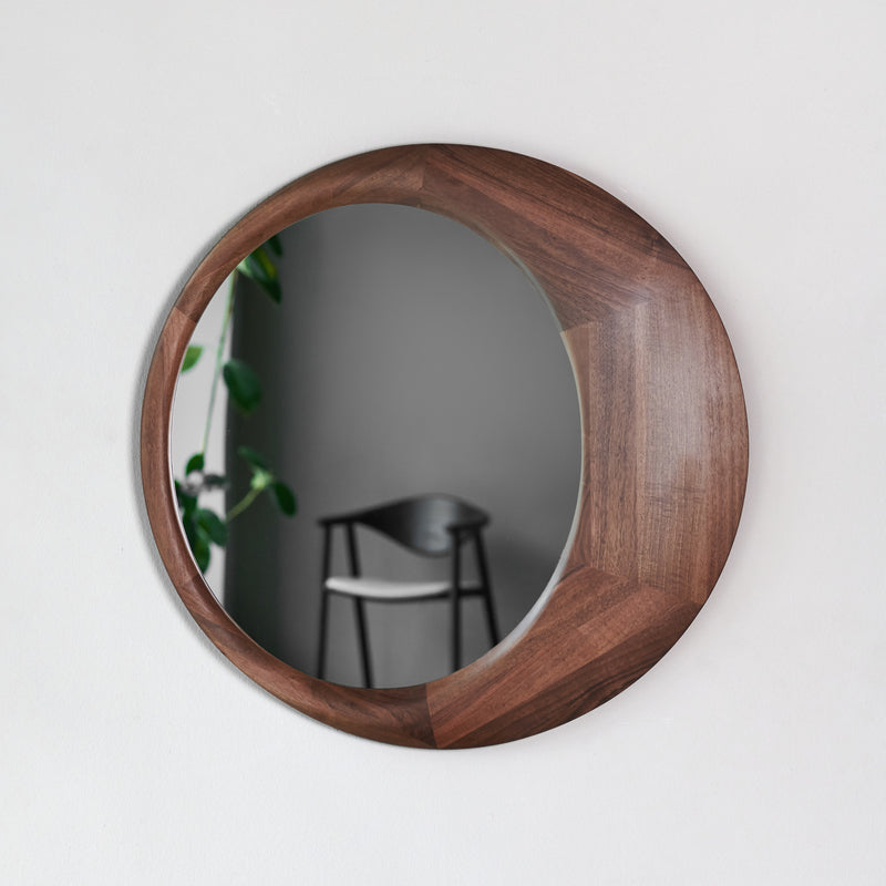 The Lana Mirror by Artisan features a solid wood frame and reflects a room with a black chair, leafy green plant, and light gray wall. Its crafted from eco-friendly materials, capturing serene surroundings.