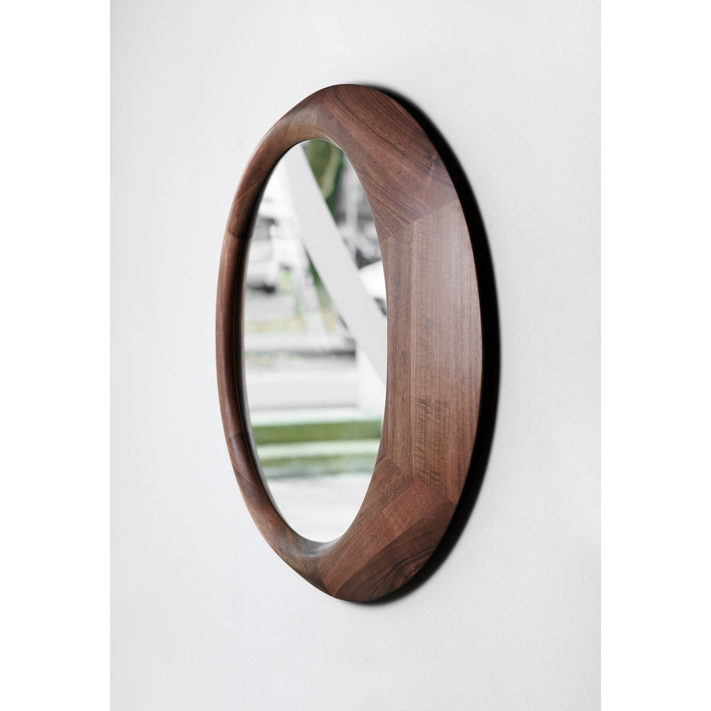 The Lana Mirror by Artisan features a thick, dark solid wood frame finished with natural oils, hanging on a white wall. Its round design reflects a blurred outdoor scene with greenery and a sidewalk, combining elegance and sustainability.