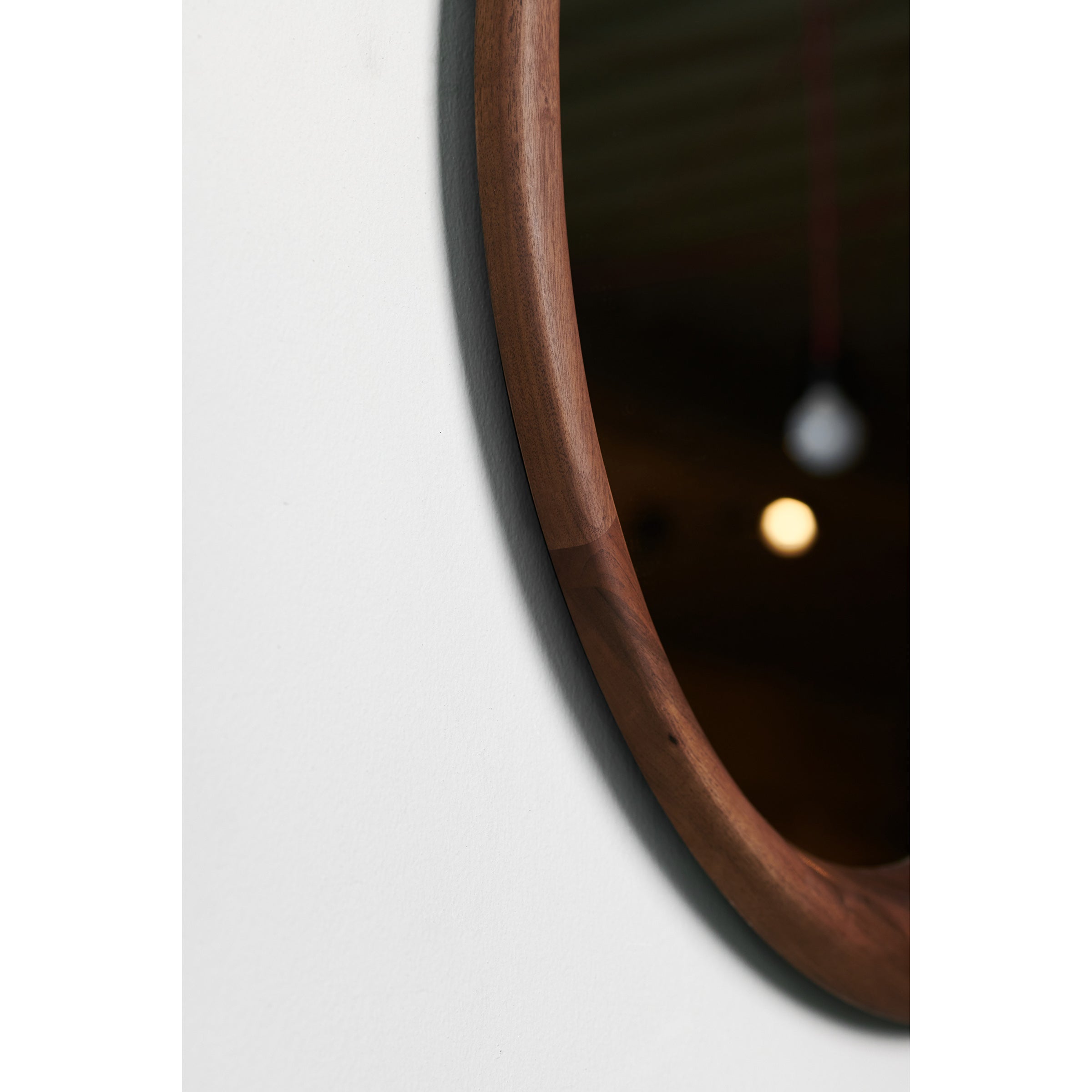 Close-up of the Artisan Lana Mirror with a solid wood frame against a white wall. A blurred light bulbs reflection creates a soft glow, emphasizing the beauty of its environmentally-friendly materials.