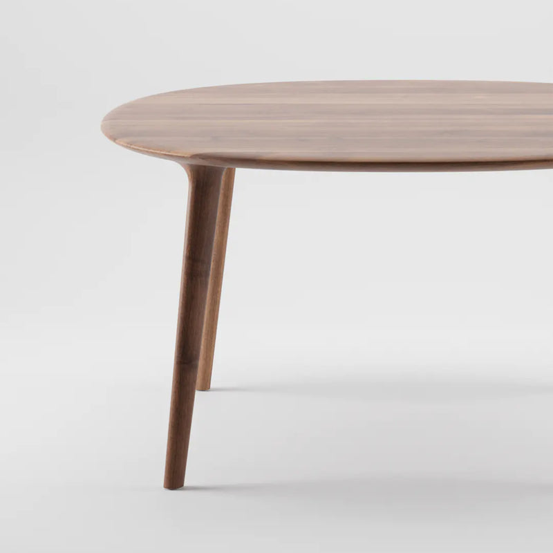 The LUC Table Round - Sale by Artisan showcases a minimalist design with smooth edges and slender legs against a light grey background, revealing only a portion of the tabletop and two legs with natural, polished wood finishes.