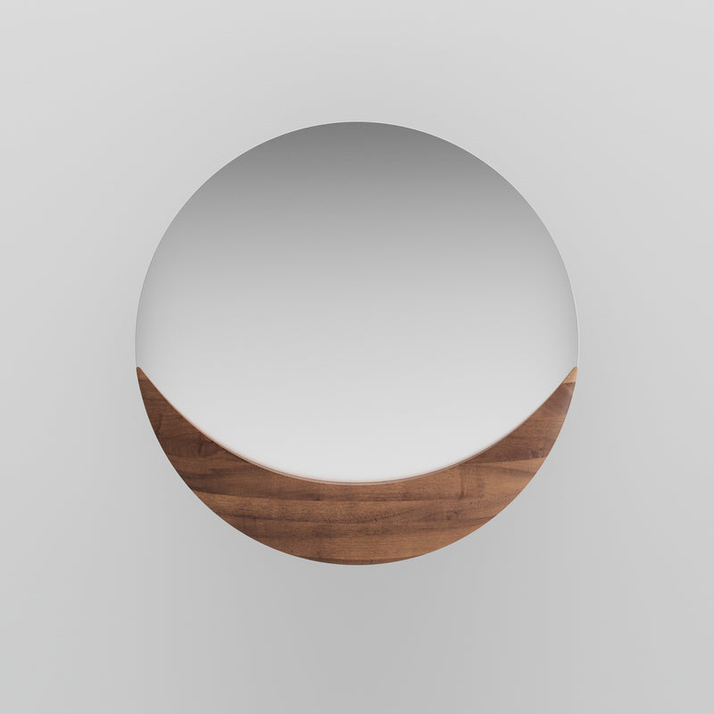 The Artisan Luna Mirror features a round design with the bottom half framed in smooth, solid wood against a plain white wall, offering an eco-friendly and minimalist aesthetic.