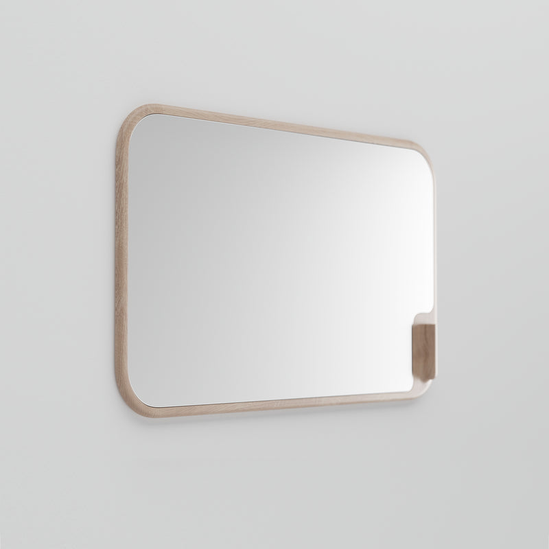 The MUSE Mirror by Artisan is a rectangular mirror with rounded corners, mounted on a light grey wall. Made from solid wood and eco-friendly materials, its light wooden frame pairs beautifully with a small geometric wooden shelf on the right side.