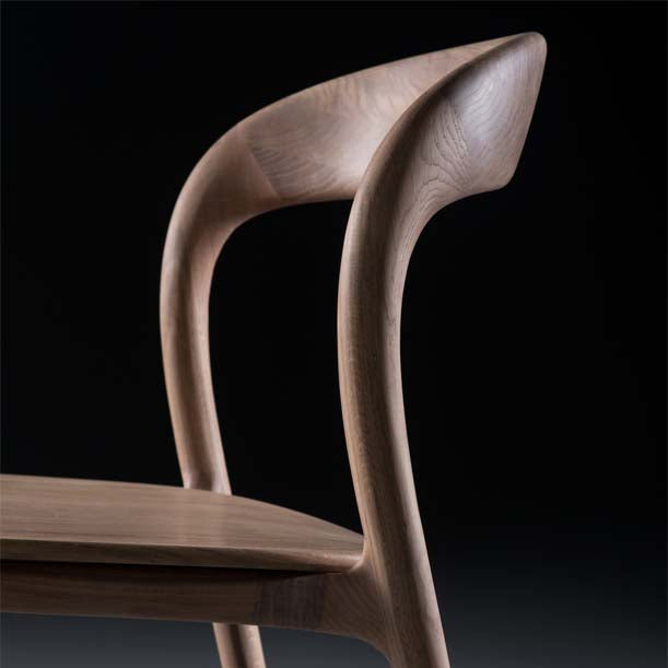 Close-up of the NEVA light chair by Artisan, showcasing an elegant wooden design with a smooth, curved backrest and seat. Visible wood grain highlights its craftsmanship, while CNC carving enhances beauty. The dark background emphasizes the exquisite chair from the Neva collection—now on sale.