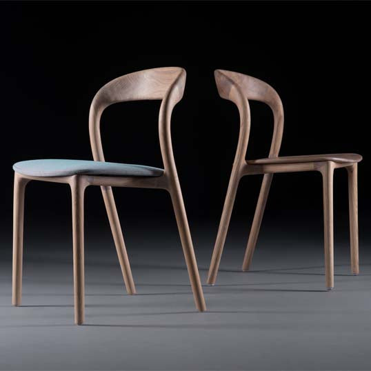 Two NEVA light chairs by Artisan, with curved backs and upholstered seats, are placed on a sleek dark surface. One chair is slightly angled, highlighting the exquisite wood craftsmanship against a black background.