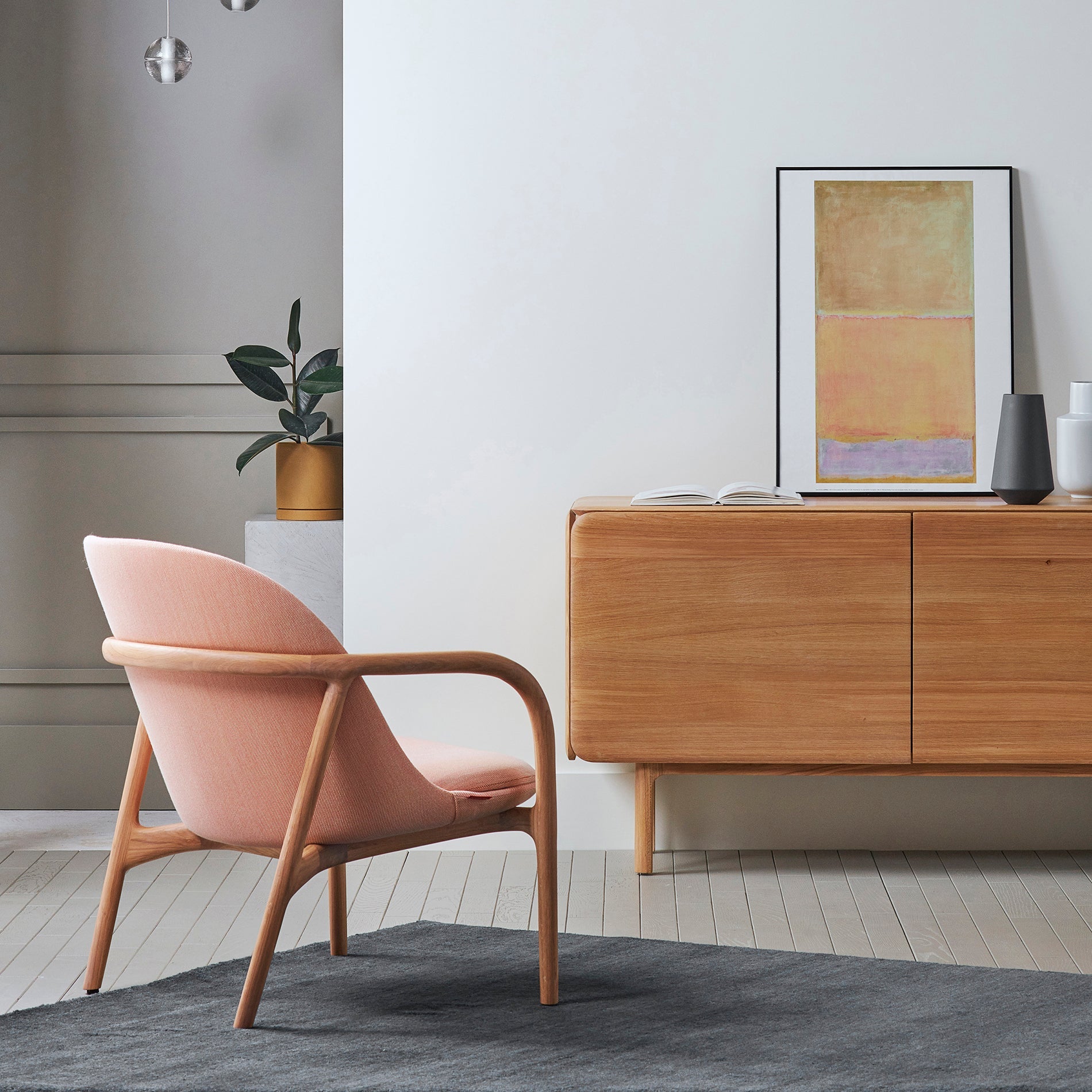 A minimalist interior features an Artisan NEVA Lounge Chair Trimmed beside a wooden sideboard with a framed abstract painting, complemented by a small potted plant and a gray rug on the light wood floor.