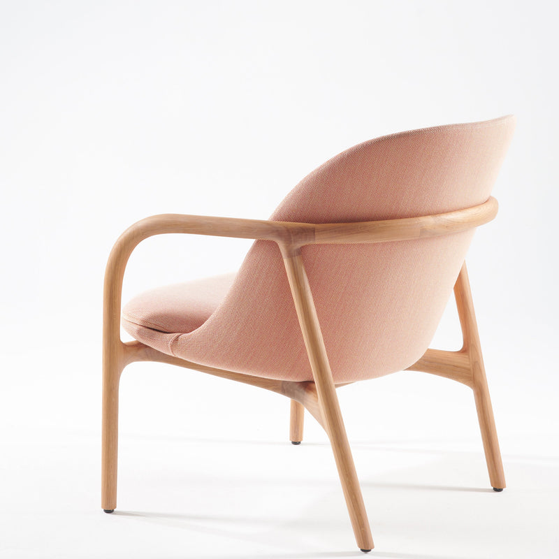 The NEVA Lounge Chair Trimmed by Artisan features a minimalist Scandinavian design with a curved backrest, light pink fabric seat, natural wood frame, and sleek rounded legs. Perfectly set against a plain white background, it exudes artisanal craftsmanship.