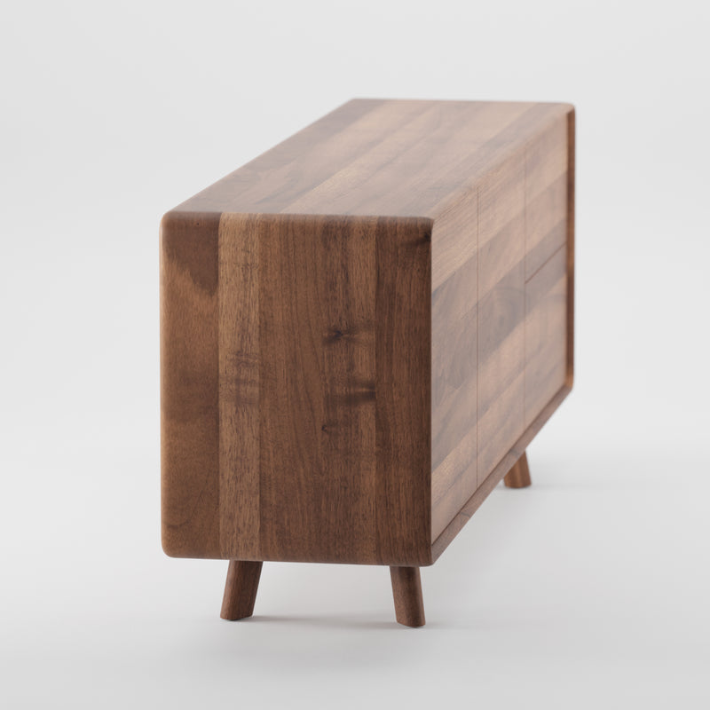 The NEVA Sideboard by Artisan showcases a minimalist design with smooth, rectangular lines, rounded edges, and short angled legs. Its rich natural finish highlights the woods grain pattern against a simple white backdrop.