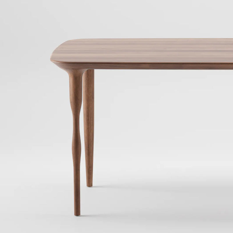 A close-up of the PASHA Table by Artisan reveals a smooth, brown surface with visible grain patterns and elegantly tapered legs featuring a unique, slightly twisted design against a plain white background.