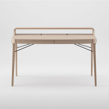 The Artisan PICARD Desk is a minimalist piece crafted from solid wood. It features four sleek legs, a raised back edge, subtle storage compartments, and black metal supports for added stability. Its plain light backdrop enhances its elegance and allows for customization.