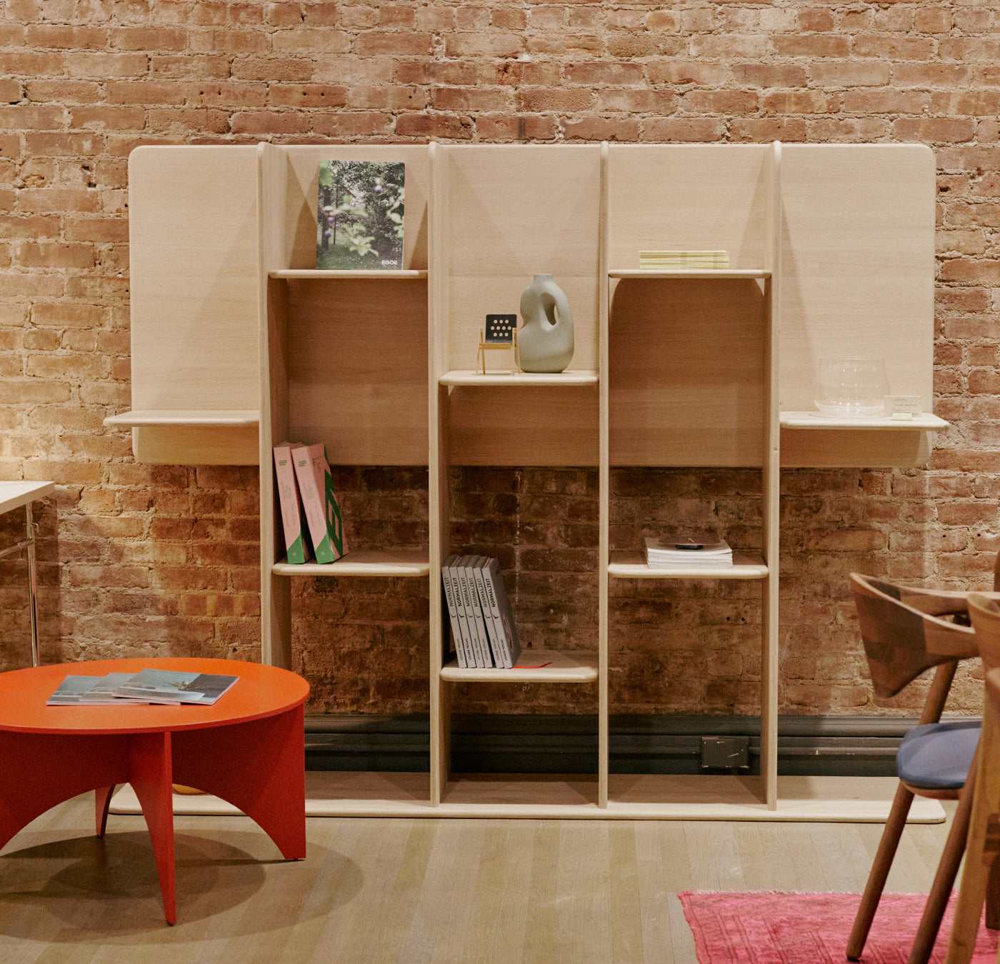 The Artisan SOFT Shelf - IVENTORY, a modern wooden unit with natural finishes, holds books, a sculpture, and decor against a brick wall. A red round table sits on the left; an eco-friendly wooden chair with cushion on the right. A pink rug peeks from underneath.